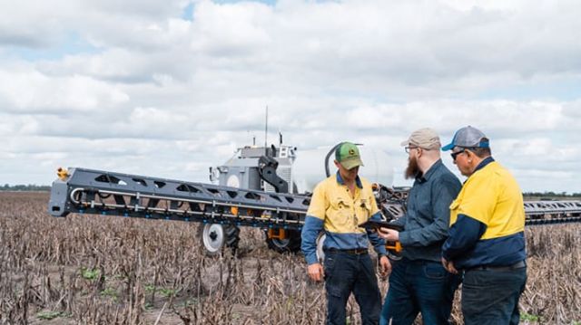 Three people in a paddock discussing a tablet beside a large spray rig.