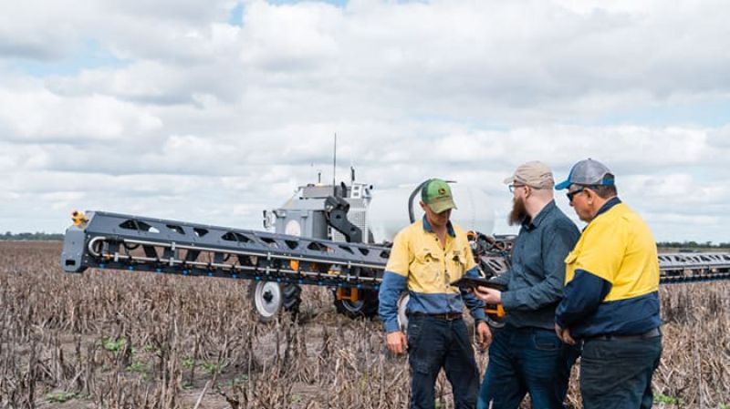 Three people in a paddock discussing a tablet beside a large spray rig.