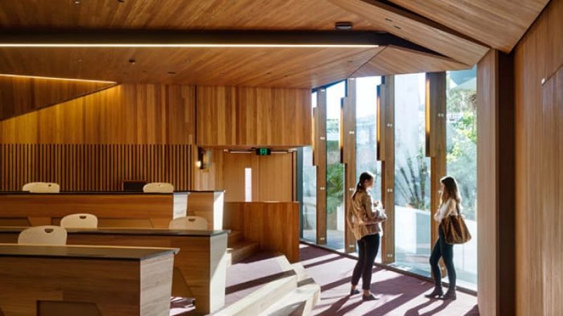 Two people talking in a timber-lined auditorium near tall windows with sunlight streaming in.