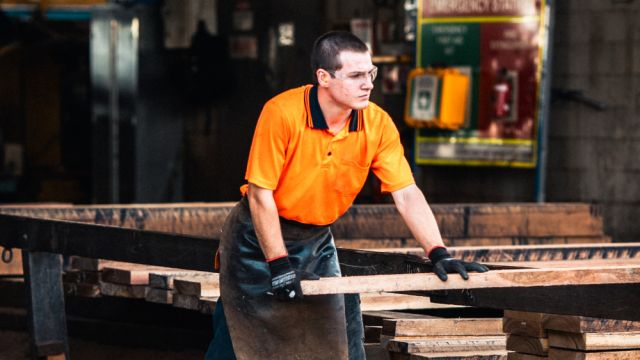Worker in hi-vis moving timber boards in an indoor processing area.