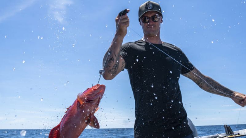 Person on a boat holding a fishing line with a red fish above the ocean, water splashing.